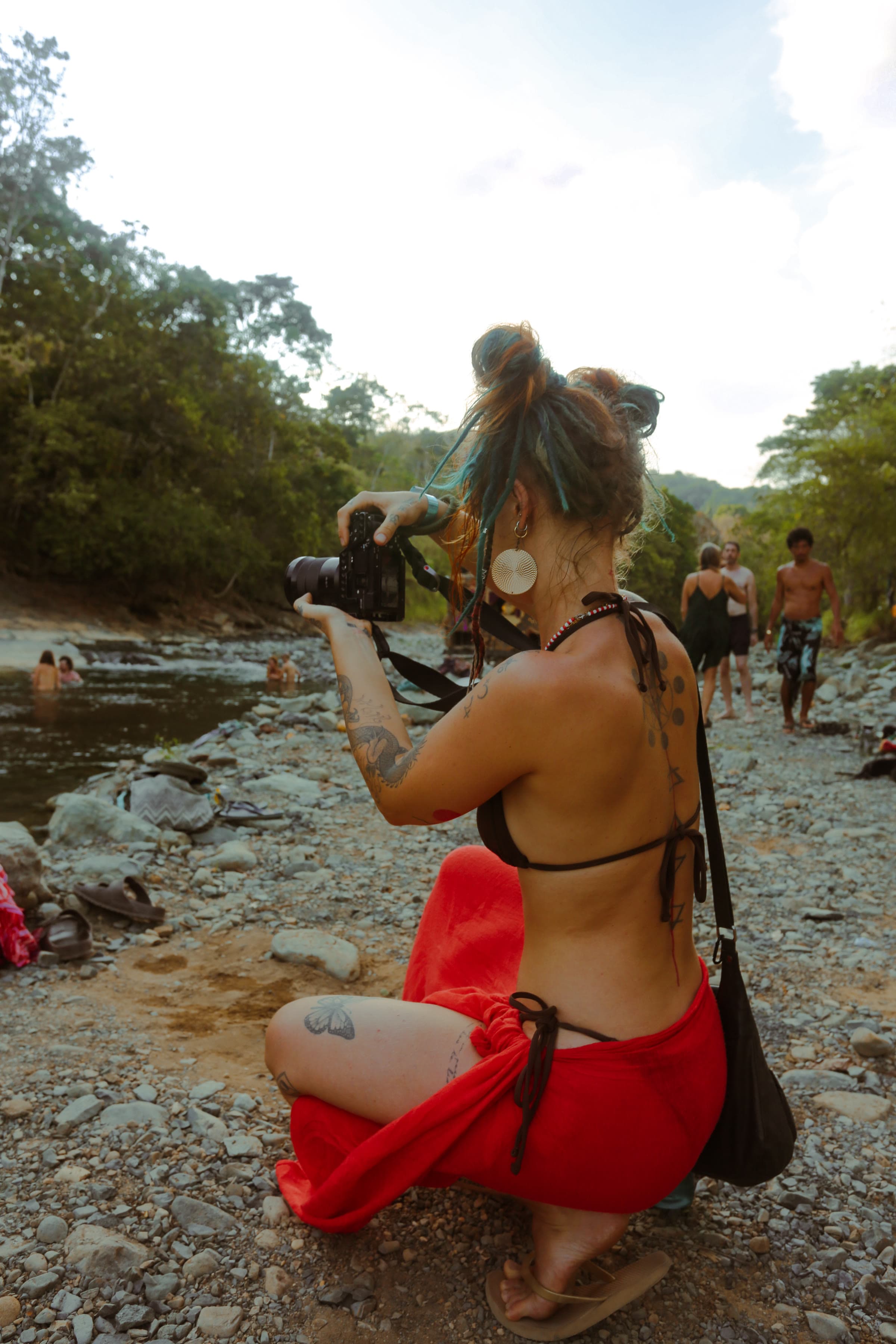 Agota by a riverbank in a red wrap with blue dreadlocks, holding a camera.