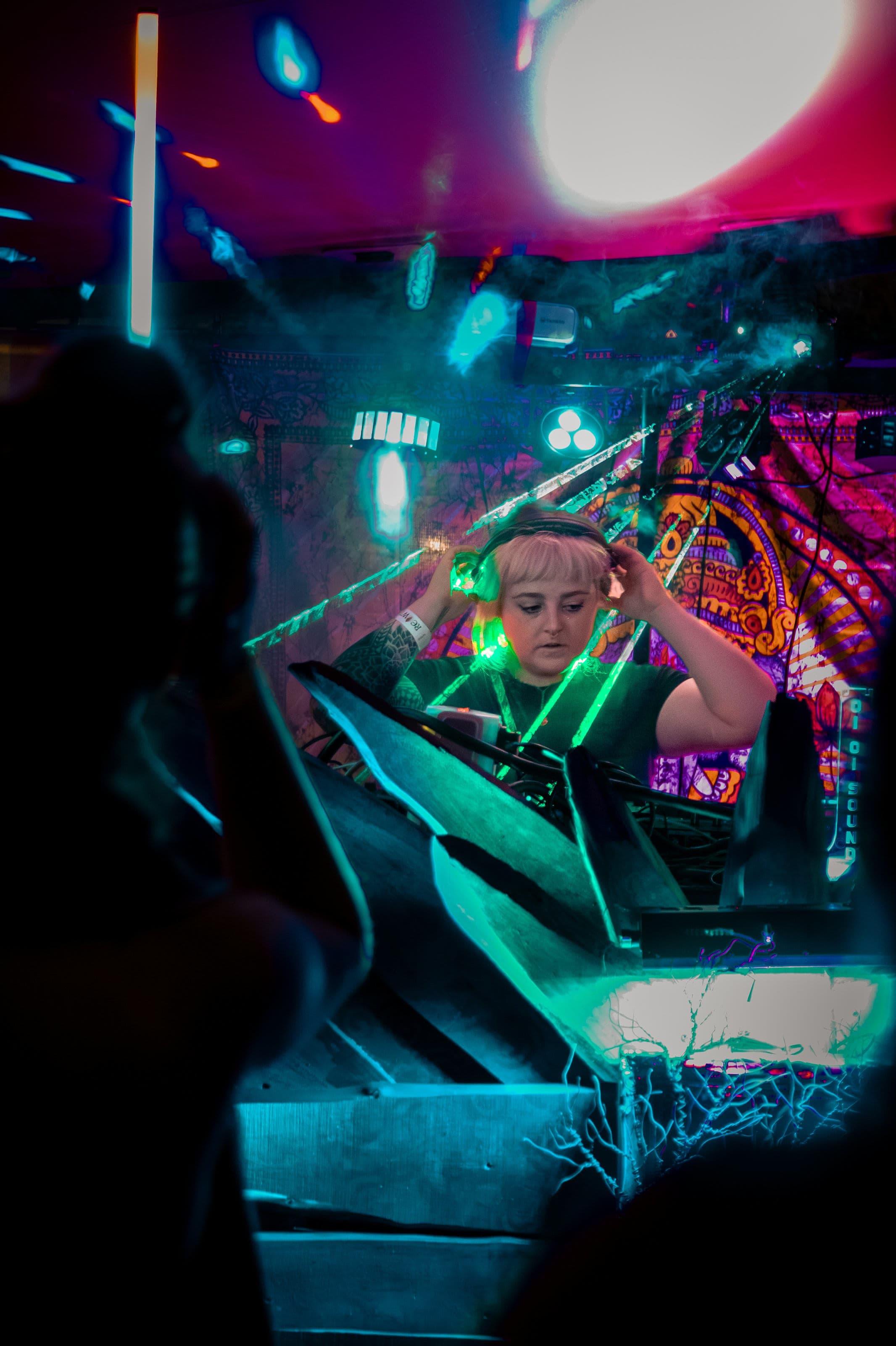 A blonde DJ adjusts her headphones behind a crystalline booth in teal light, with a psychedelic backdrop and club lasers.