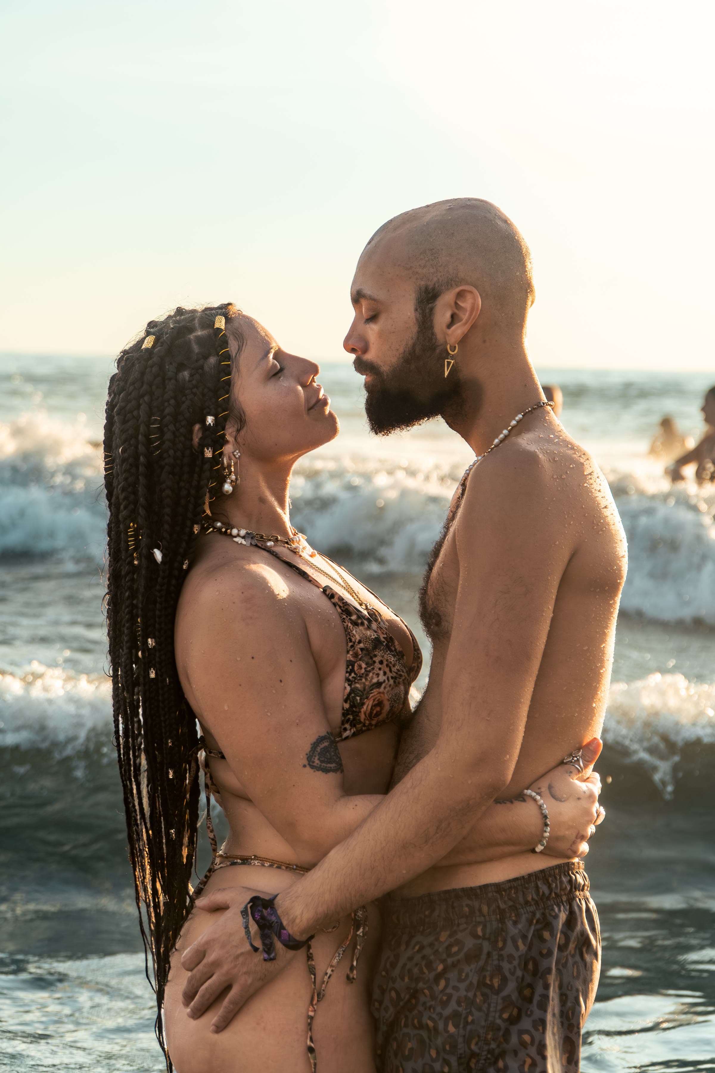 A couple stands waist-deep in golden-hour waves, eyes closed and foreheads nearly touching: she faces forward in long dark braids with gold cuffs and a leopard bikini, he is bald and bearded in matching trunks.