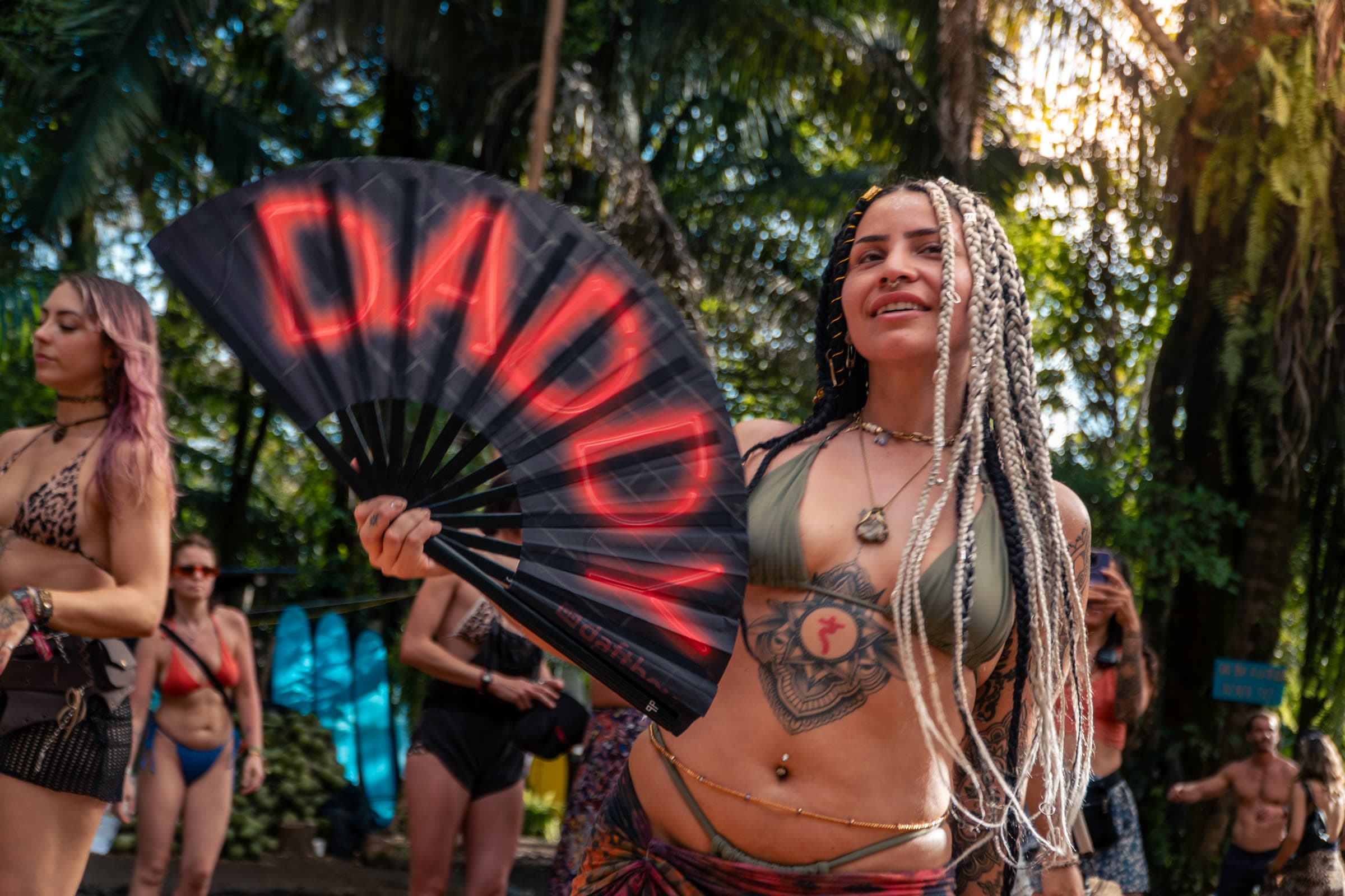 A woman with blonde and black micro-braids holds a neon-red fan that reads “DADDY,” wearing an olive bikini and a patterned wrap among tropical festival foliage.