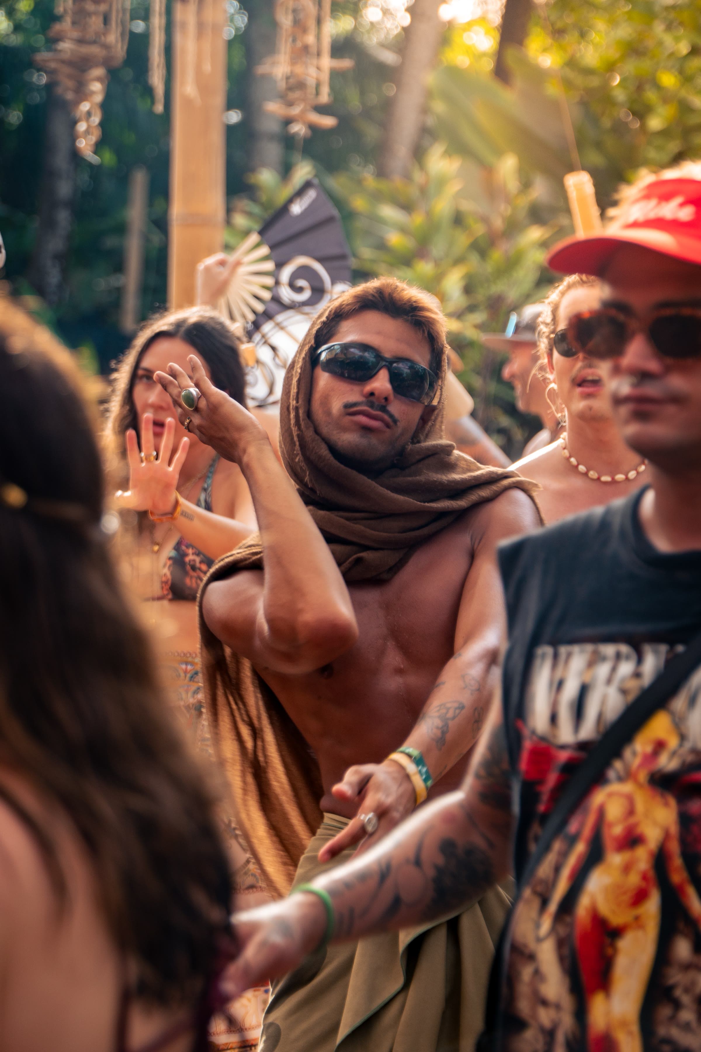 A shirtless man in sunglasses dances in a brown fabric wrap pulled over his head, golden light on a festival crowd behind him.