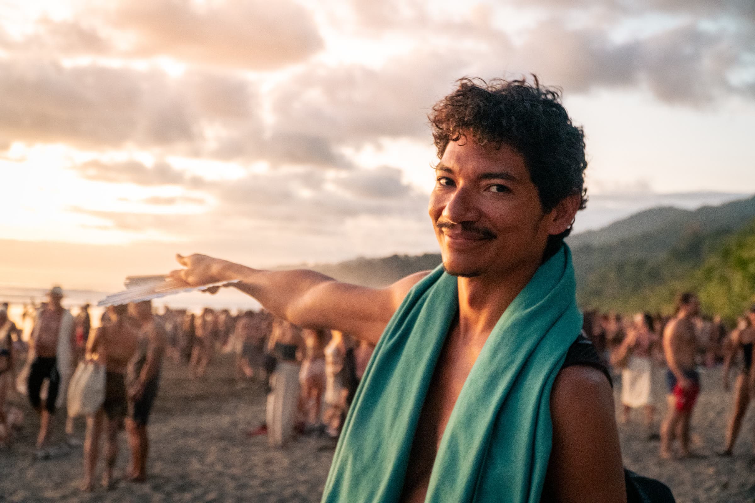 A man with a teal towel over his shoulders smiles at the camera on a beach at sunset, a crowd behind him.
