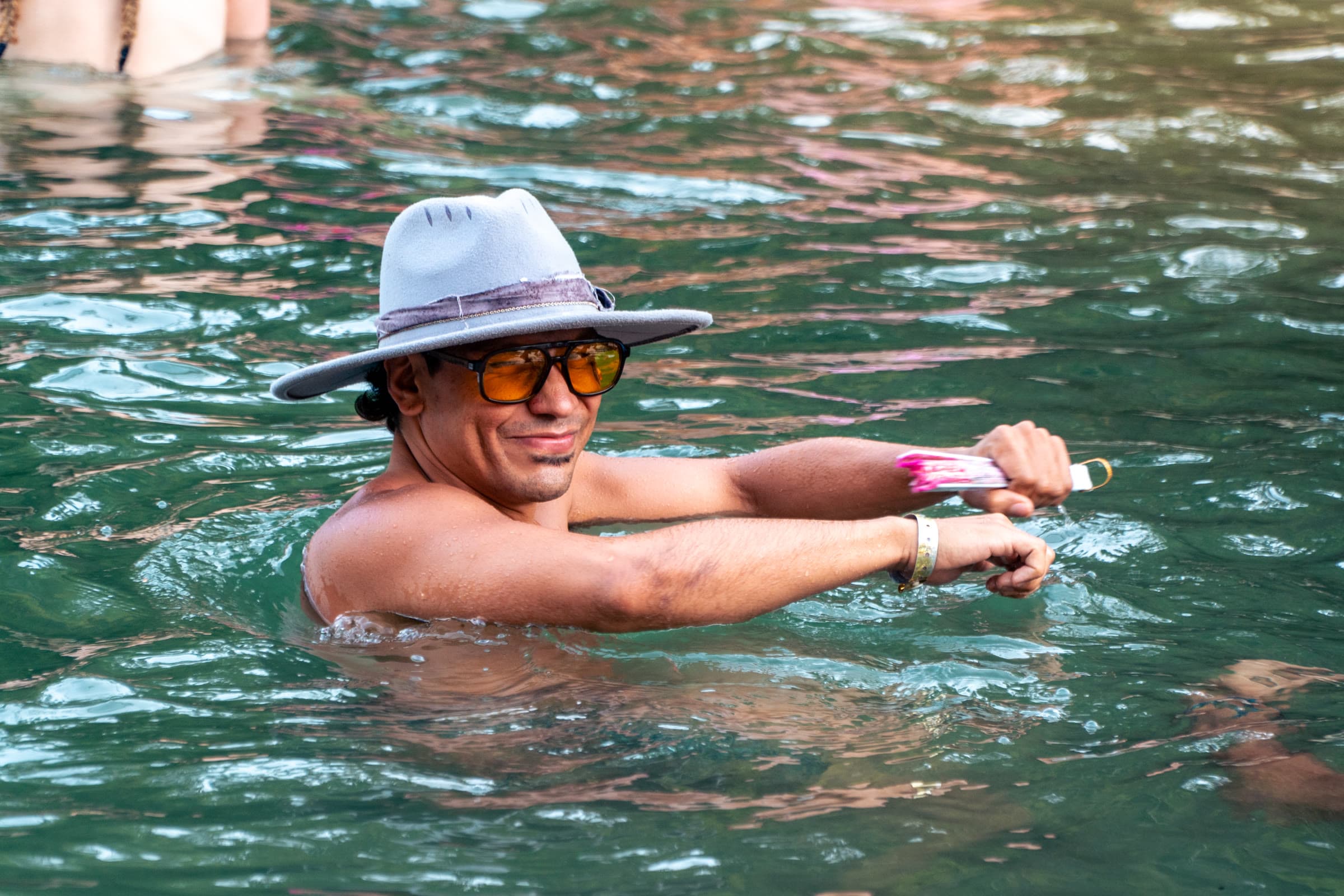 A man in a grey felt hat and orange aviator sunglasses stands in teal water holding a pink fan, with a playful smirk.