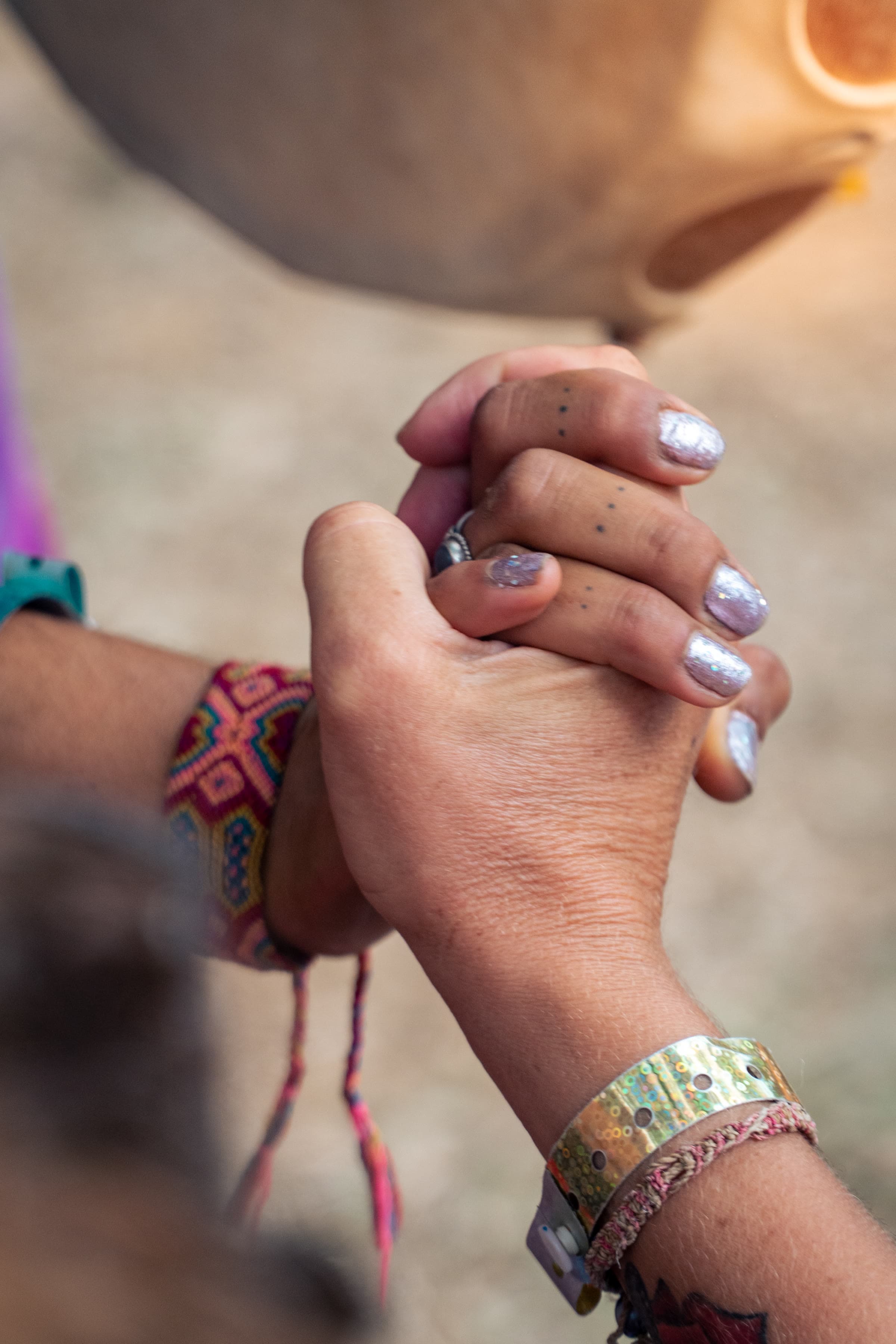 A close view of two clasped hands with festival wristbands and glitter nails, a handpan soft in the background.