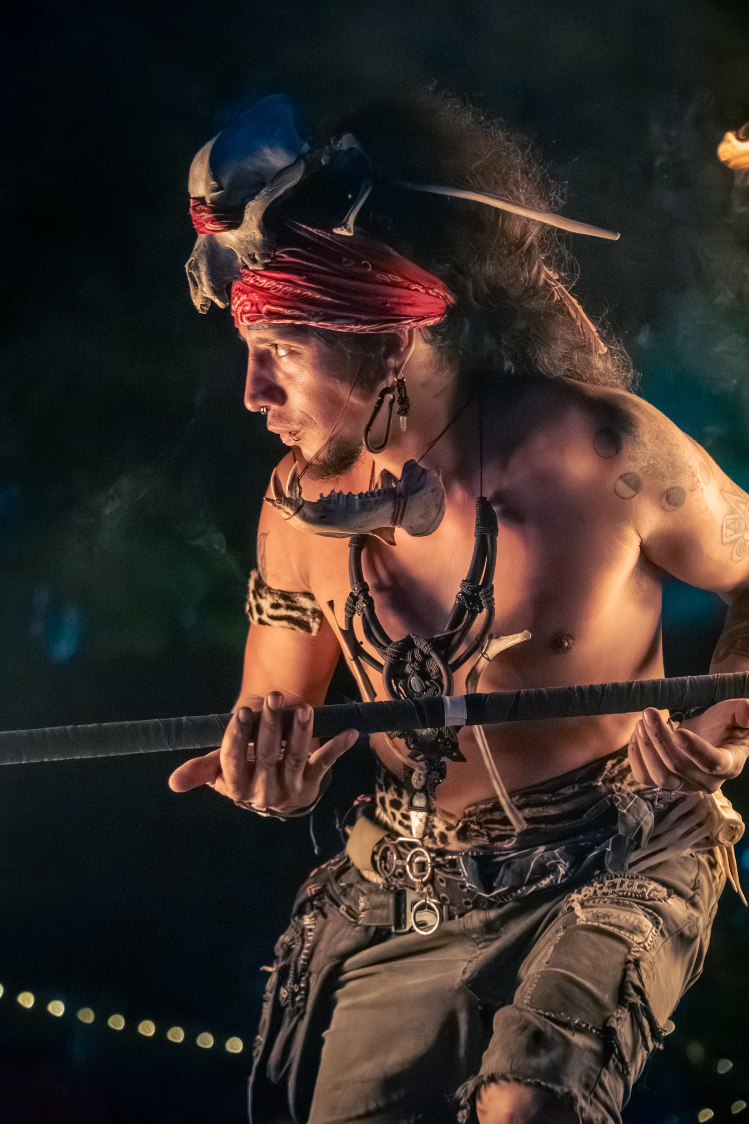 A man in a skull headdress and red bandana holds a staff during a firelit night performance.