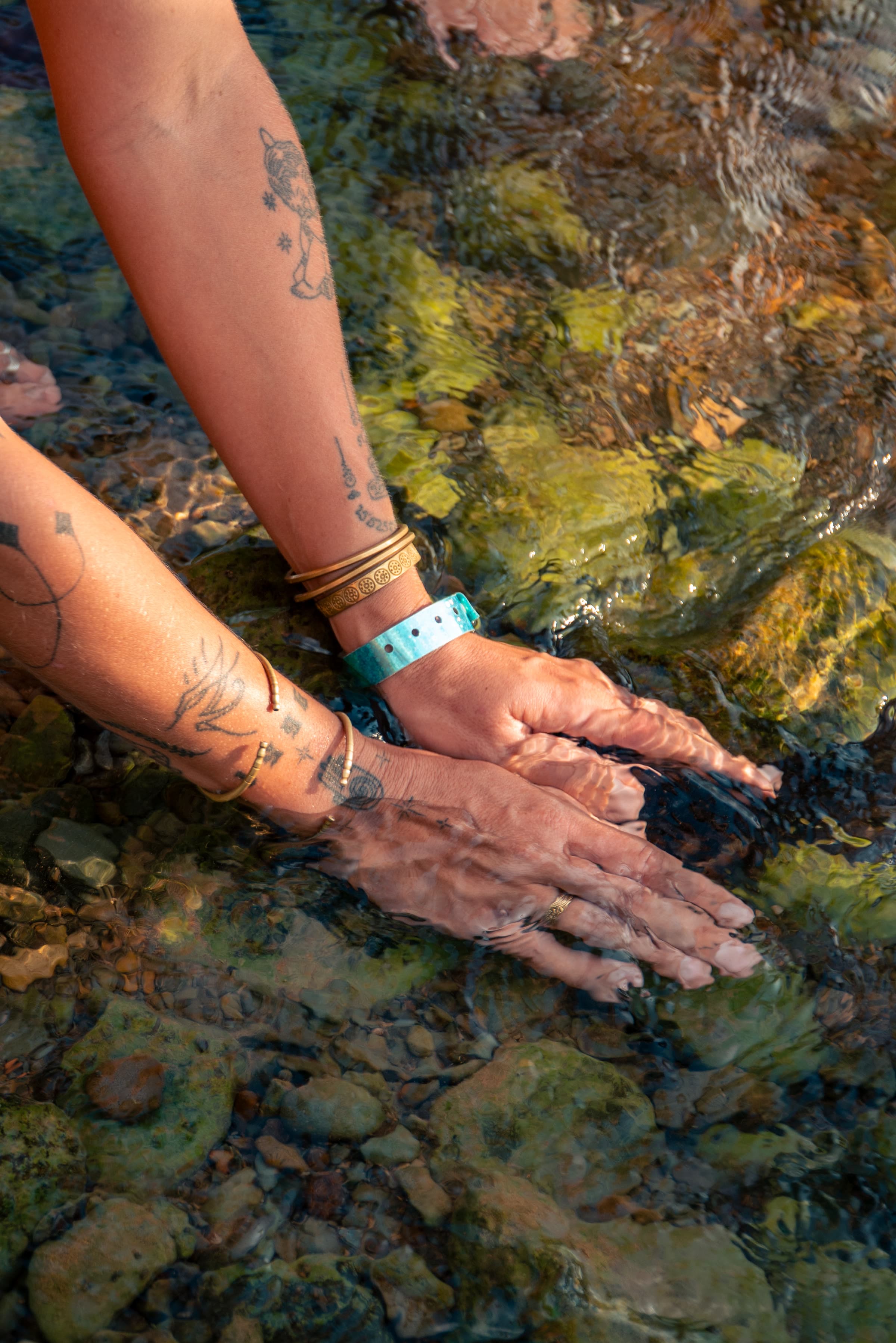 Two tattooed arms reach into clear shallow water over river rocks, gold bracelets catching the light.