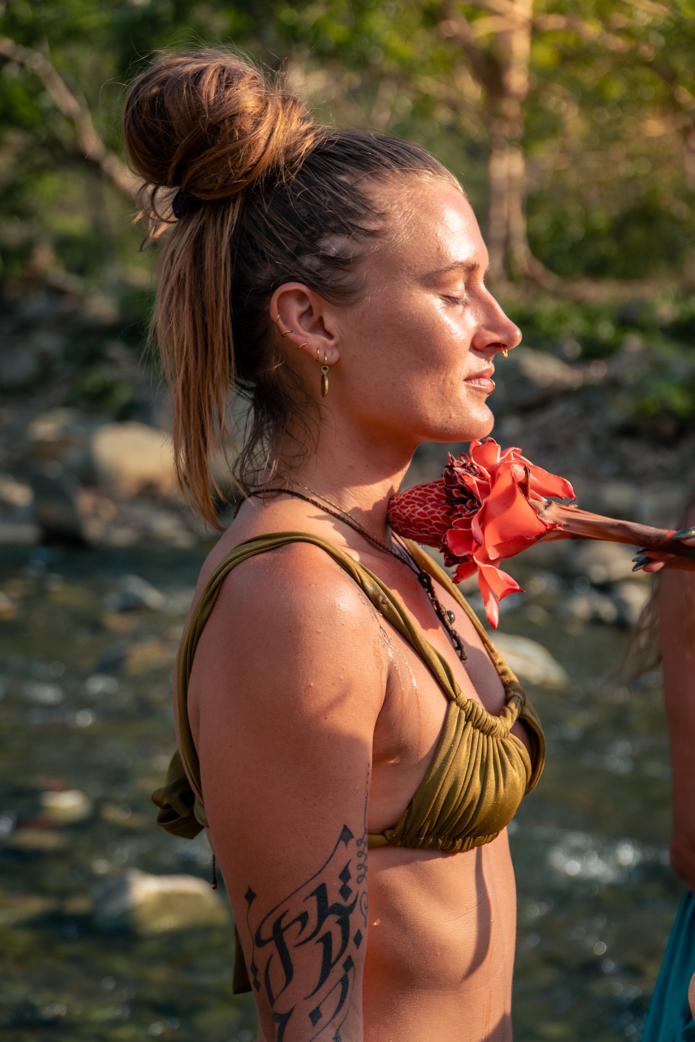 A woman in profile, eyes closed, holds a red torch ginger flower near her throat, river rocks behind her in golden hour light.