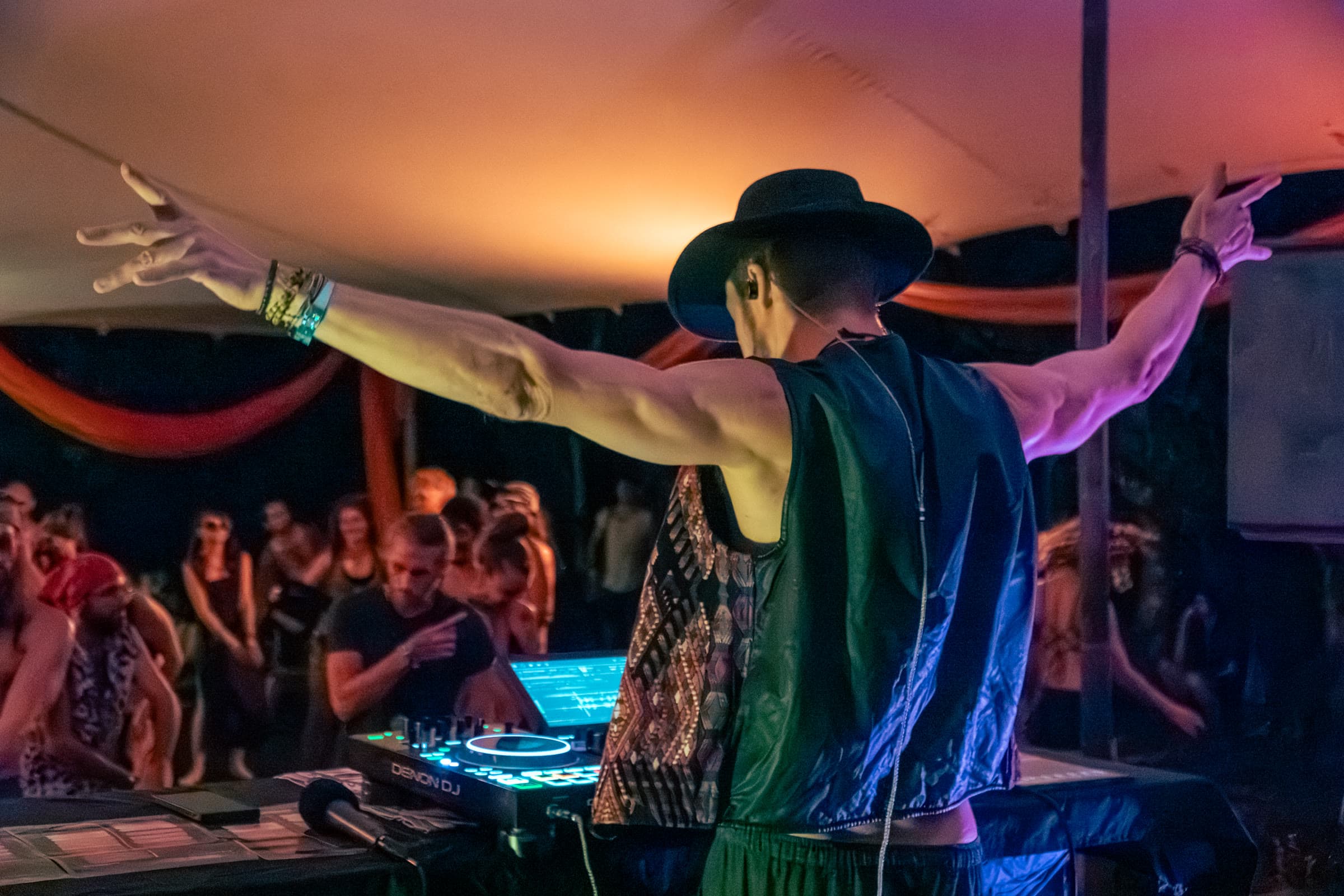 A DJ seen from behind, arms outstretched under a tent at night, a Denon deck in front and the crowd beyond.