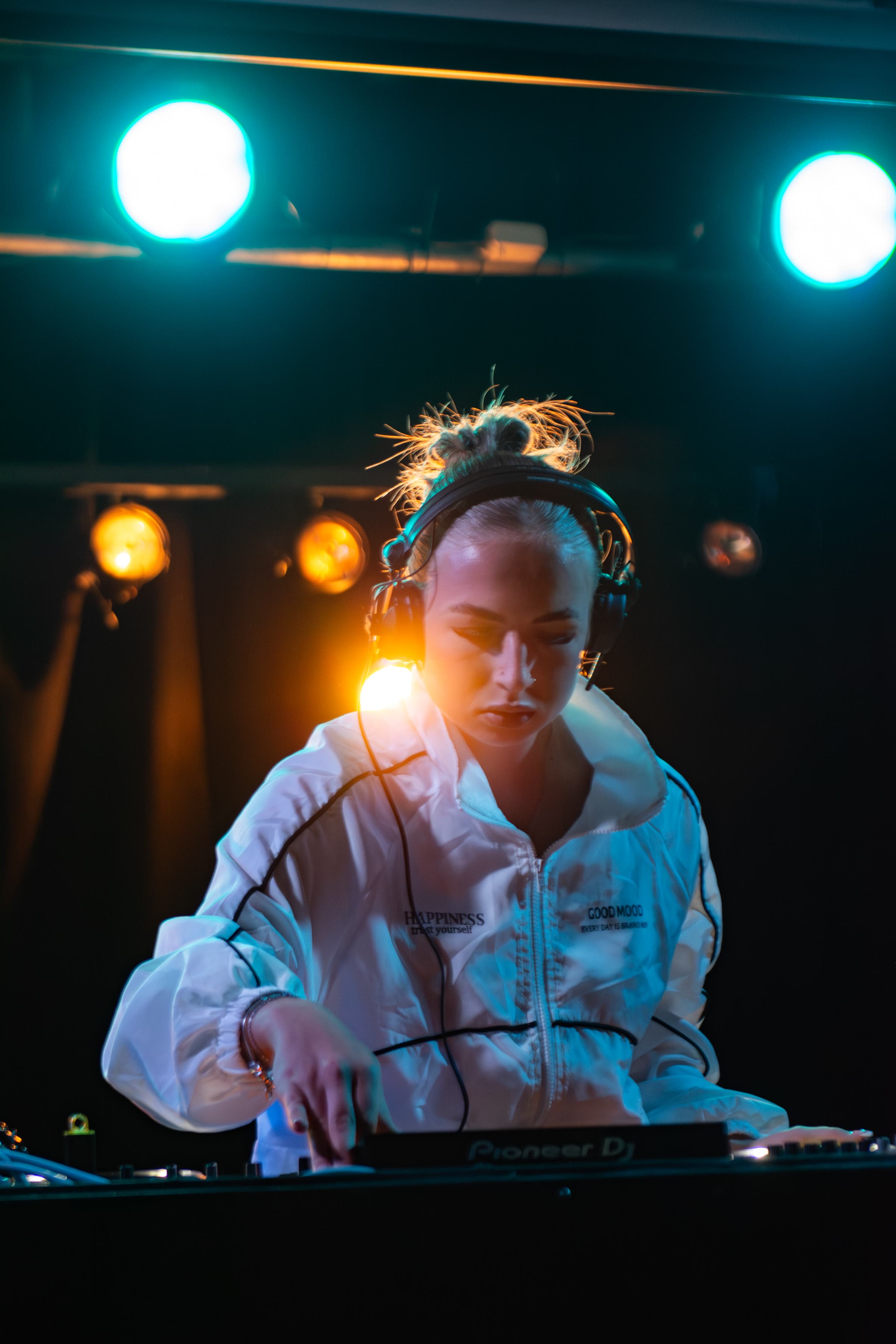 A blonde DJ in a white jacket that reads “HAPPINESS / GOOD MOOD,” wearing headphones at a Pioneer controller under cyan and amber stage lights.
