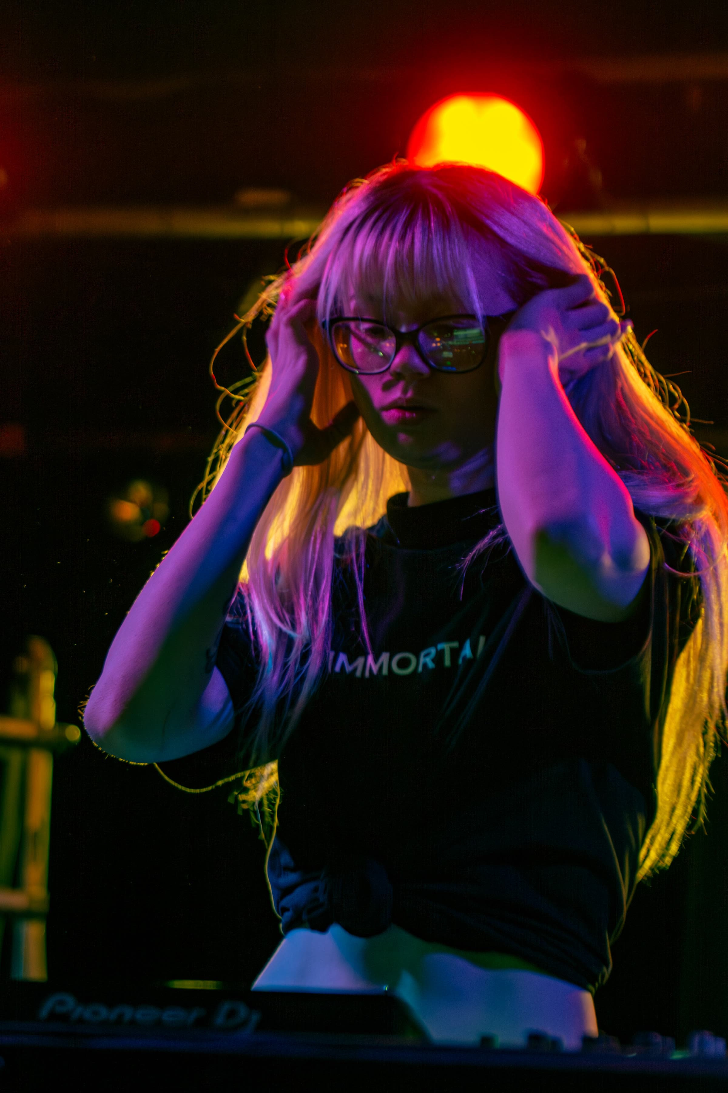 A blonde DJ with bangs and black glasses, in a knotted tee that reads “IMMORTAL,” hands in her hair, red and orange light behind a Pioneer deck.