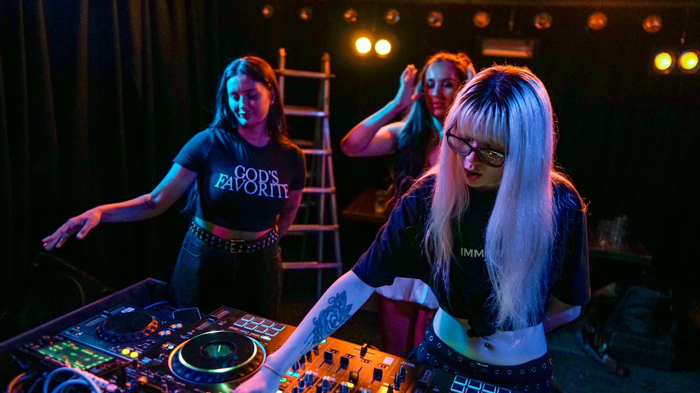 A platinum-blonde DJ with glasses at the CDJs and mixer, a woman in a “GOD’S FAVORITE” tee beside her and a dancer behind, in blue, purple, and orange club light.