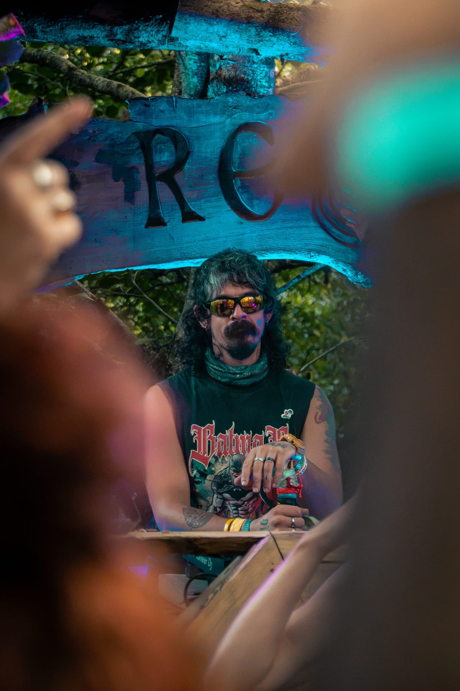 A man with dark wavy hair, a mustache and goatee, and yellow-tint sunglasses stands at an outdoor DJ booth in a green Batman sleeveless tee, shot through the crowd, a wooden “REC” sign glowing blue.