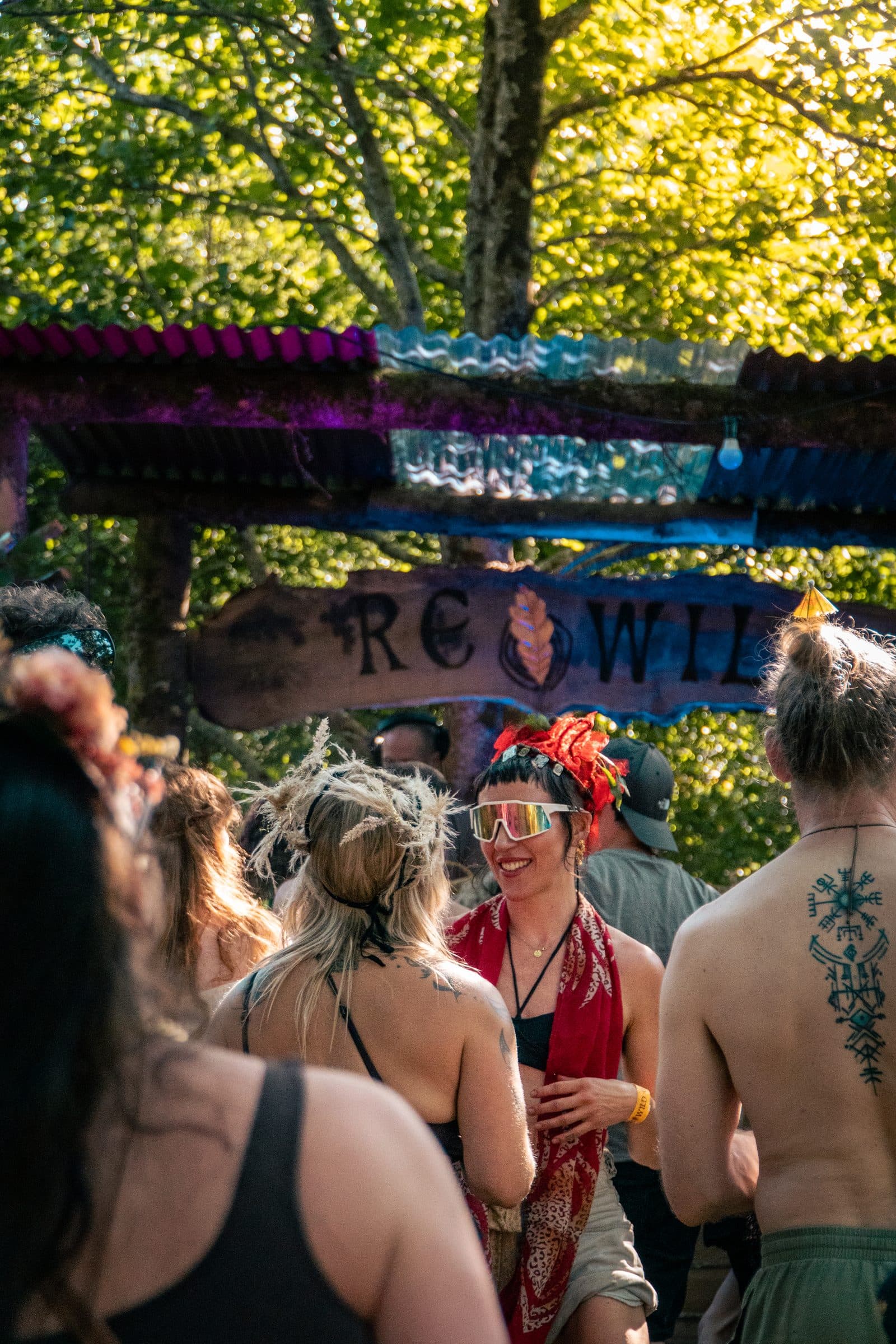An outdoor festival group under a wooden sign that reads “REWILD,” warm sun through the trees: a smiling woman in a red headpiece and white sunglasses, a shirtless man with a spine tattoo, and a man in an olive baseball cap.