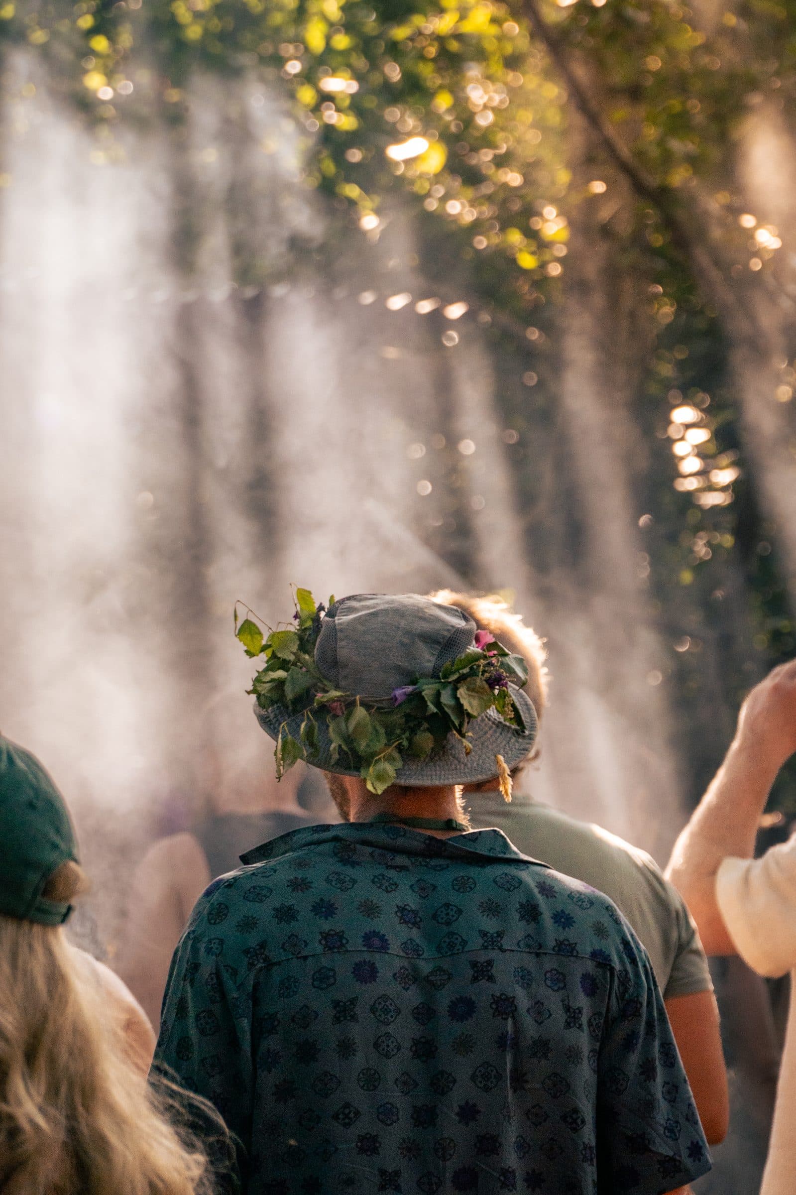 A person seen from behind in a grey bucket hat trimmed with green leaves and purple and pink wildflowers, wearing a patterned blue shirt, sunbeams through misty trees.