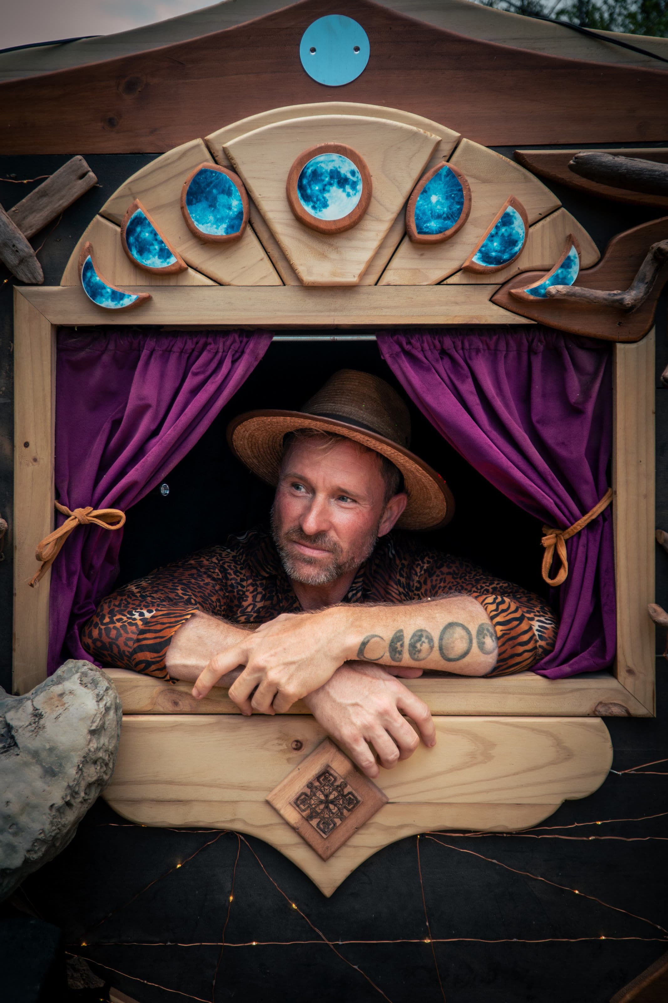 A man in a straw hat and animal-print shirt leans on a wooden booth window framed by glowing blue moon phases and purple curtains, fairy lights below.