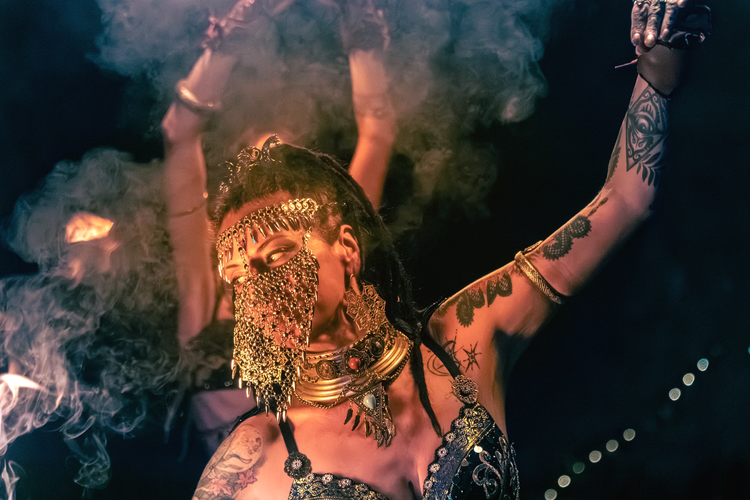 A woman in a gold chain-mail veil and crescent headdress dances in firelit smoke, tribal-fusion jewelry and tattoos along her arm.
