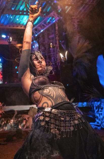 A fire performer with a wire headpiece raises a flaming prop, teal stage lights and smoke around her, a coin belt at her waist.