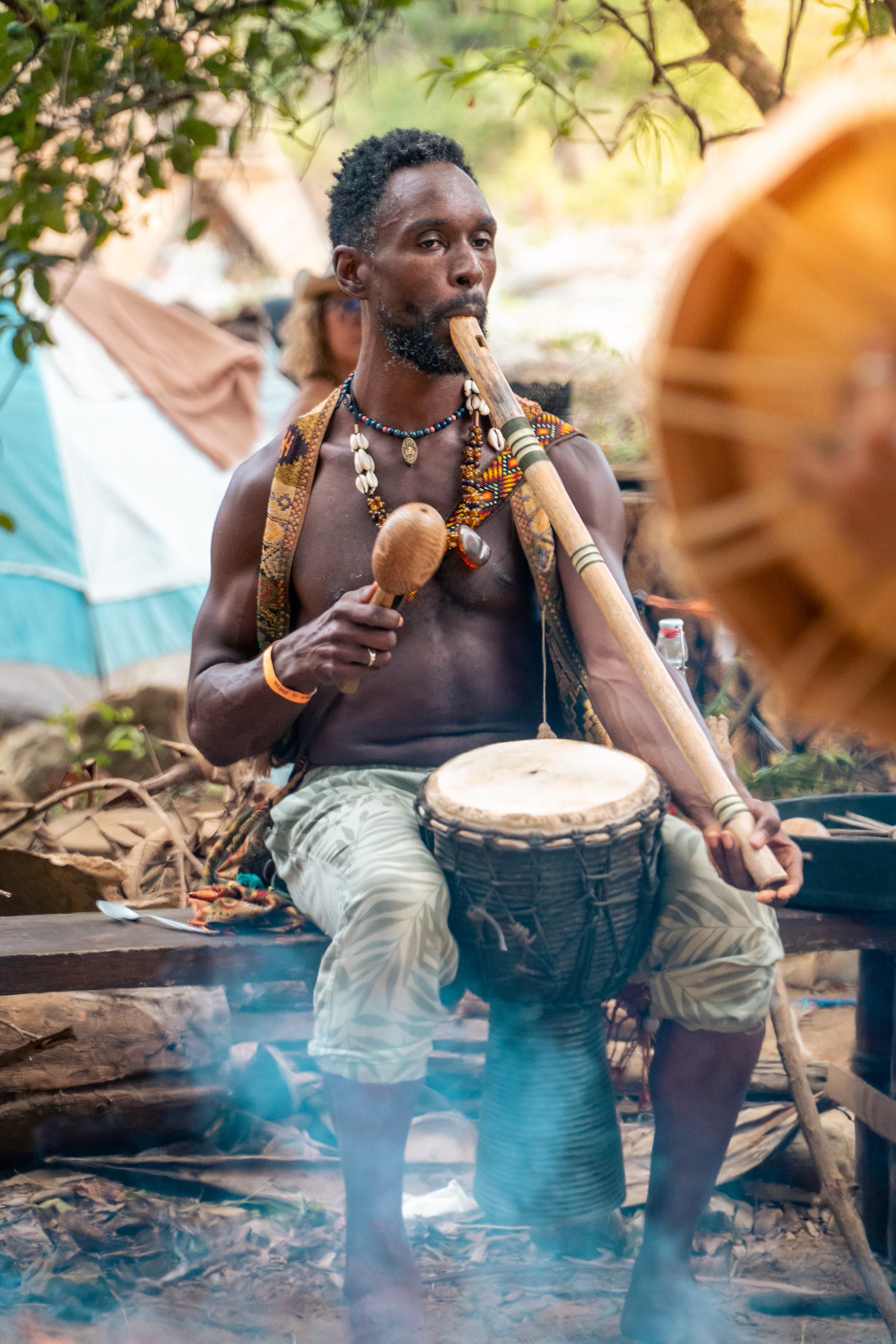 A musician outdoors plays a wooden flute beside a djembe, a maraca in the other hand, smoke drifting past.