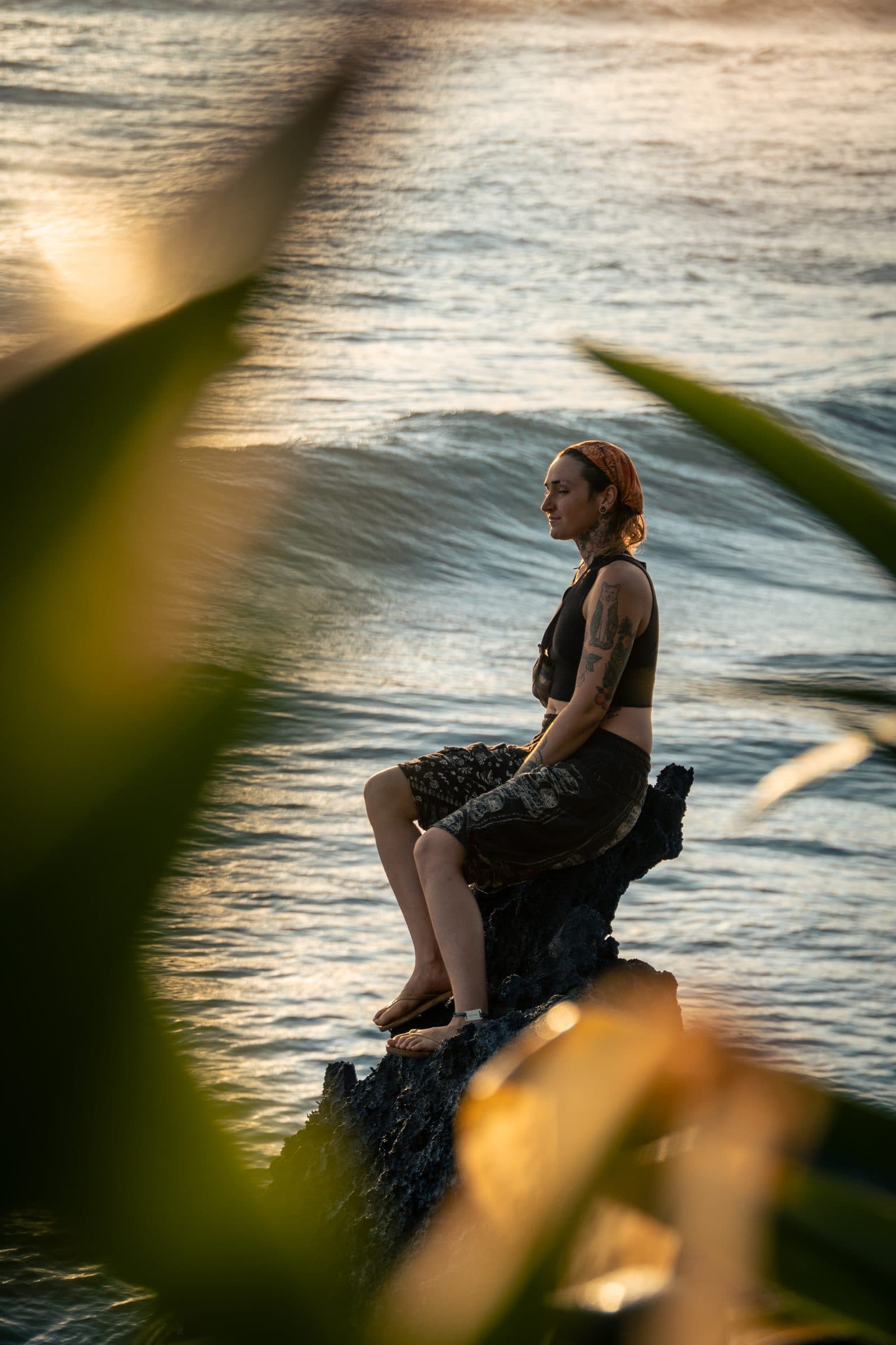 A person sits in meditation on a dark ocean rock at golden hour, an orange-red bandana over their hair, a black crop top, framed by out-of-focus green leaves.