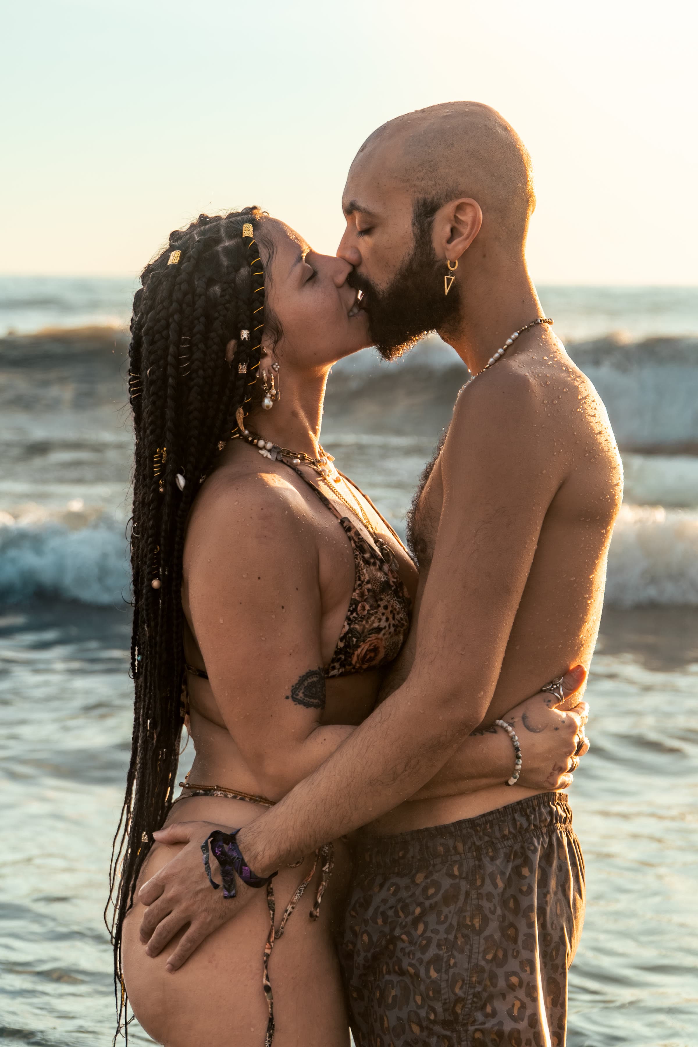 A couple kissing in the ocean at golden hour: she wears long dark braids with gold cuffs and a leopard bikini, he is bald and bearded in matching leopard trunks.