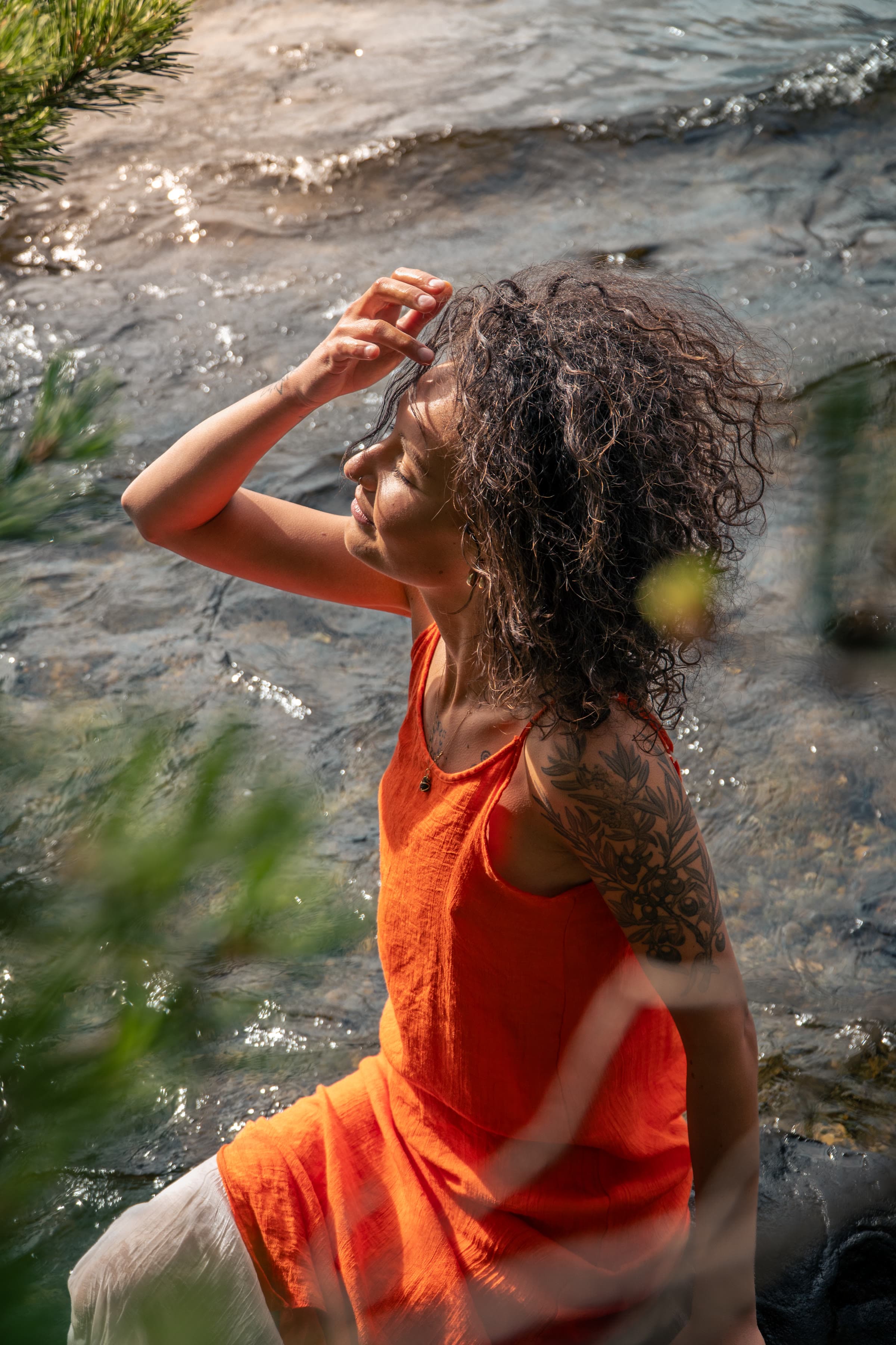 A woman with wet dark hair and closed eyes stands in clear shallow water, wearing a terracotta top, a botanical tattoo along her arm, pine needles in the foreground.