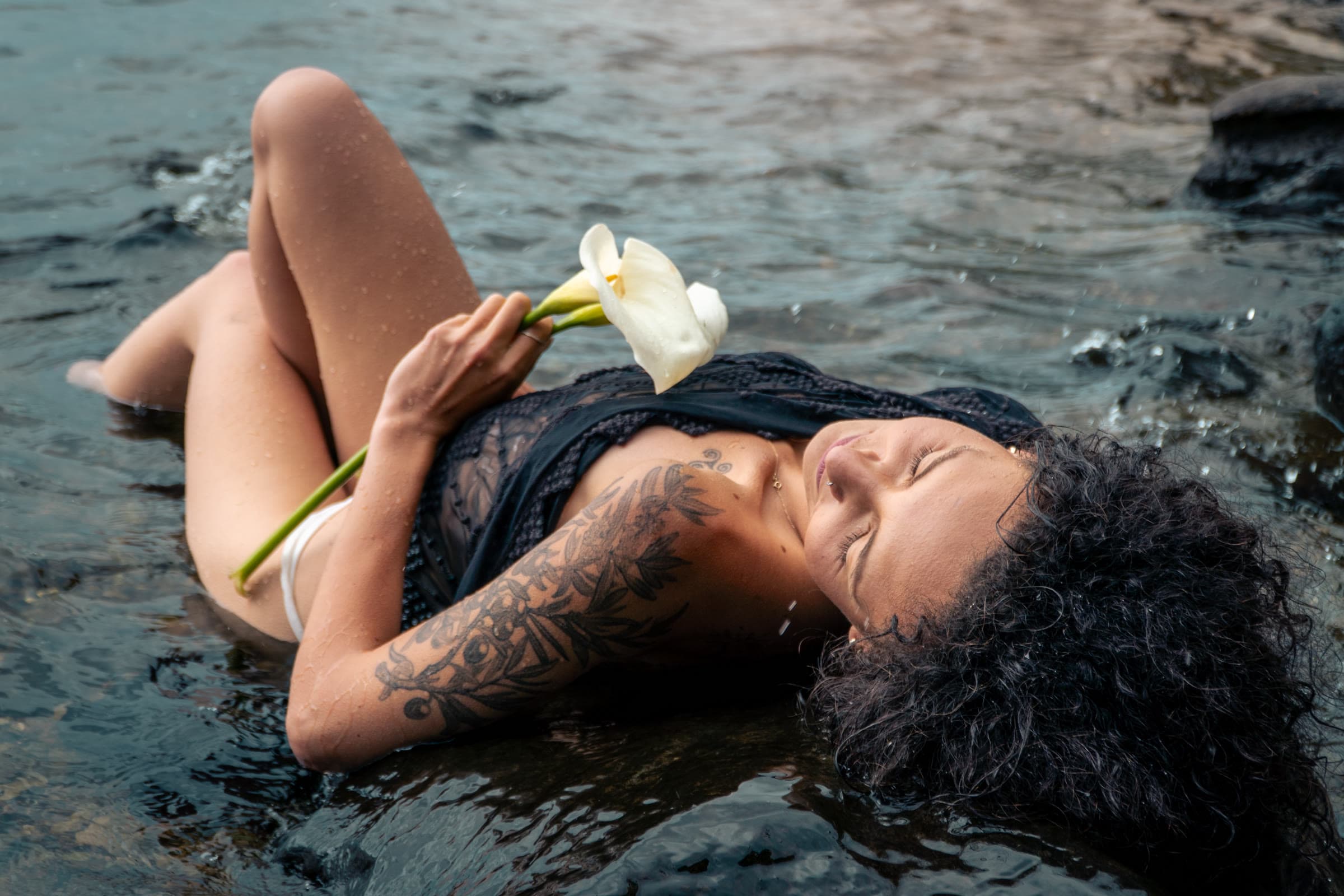 A woman lies in dark shallow water holding white calla lilies, wearing a black lace top, her wet hair spread around her.