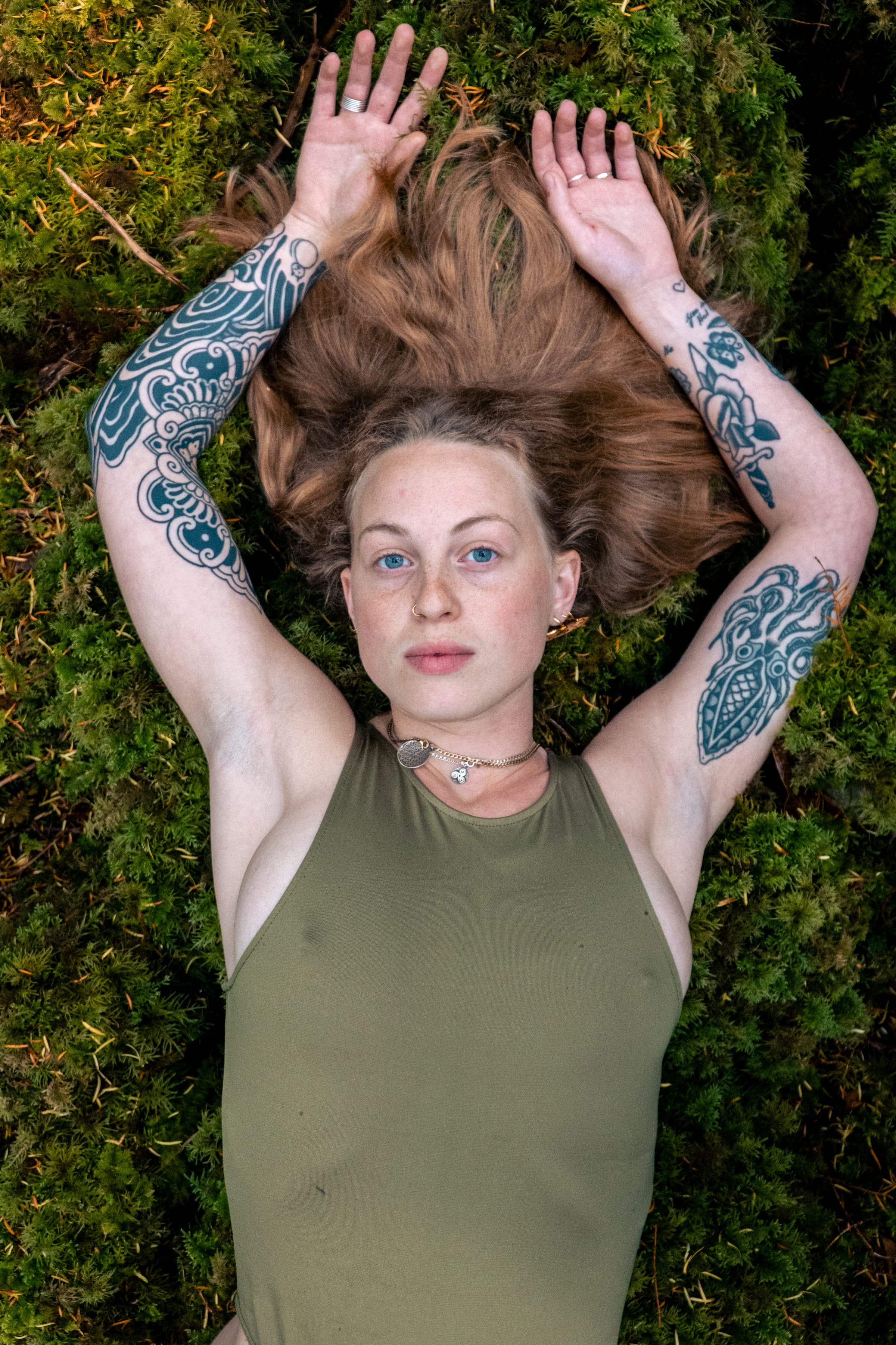 A blonde woman in an olive bodysuit lies on bright green moss and looks toward the camera, tattoos running along her arms.