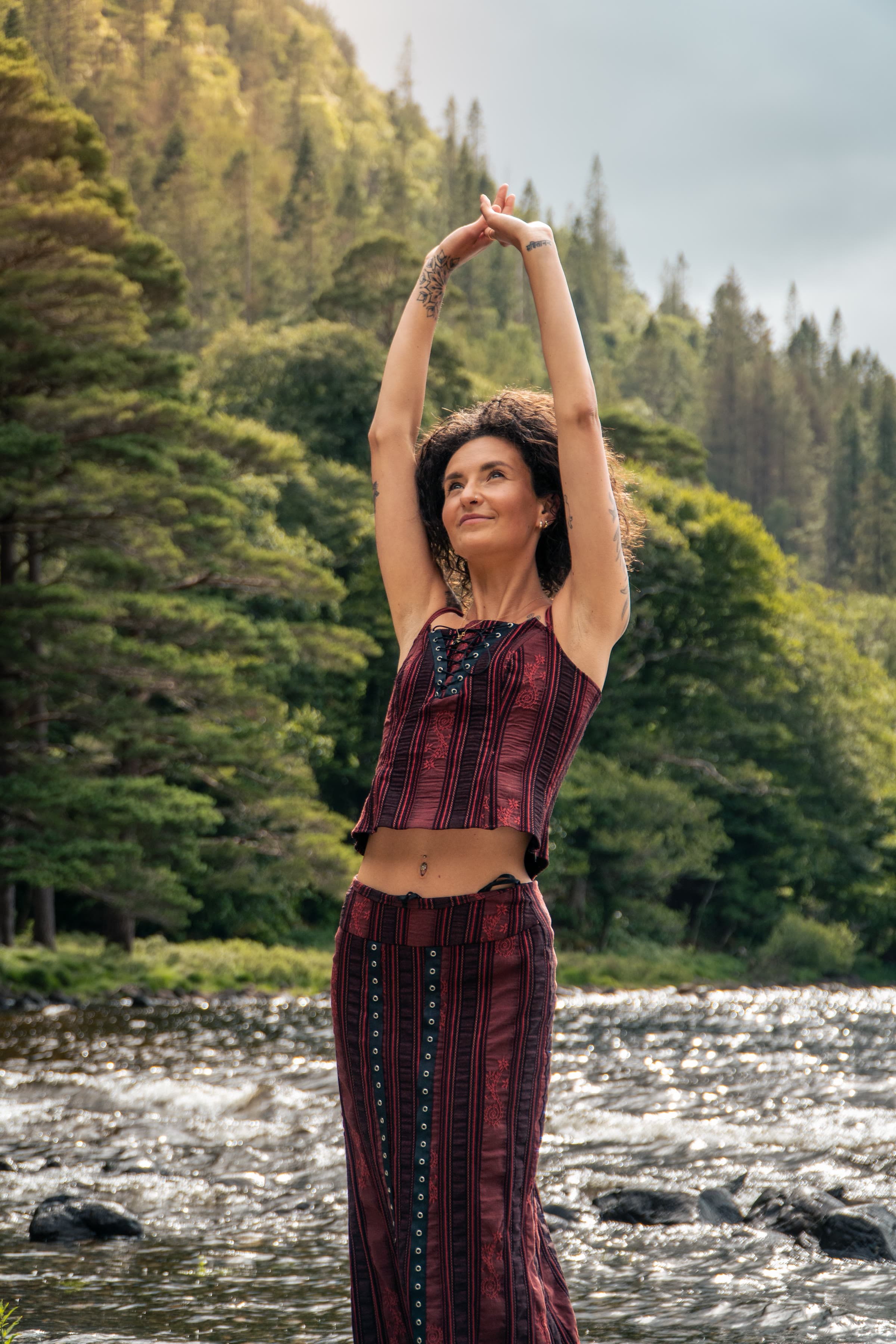 A woman with curly hair and arms raised stands beside a river in a maroon striped set, a green forested hillside behind her.