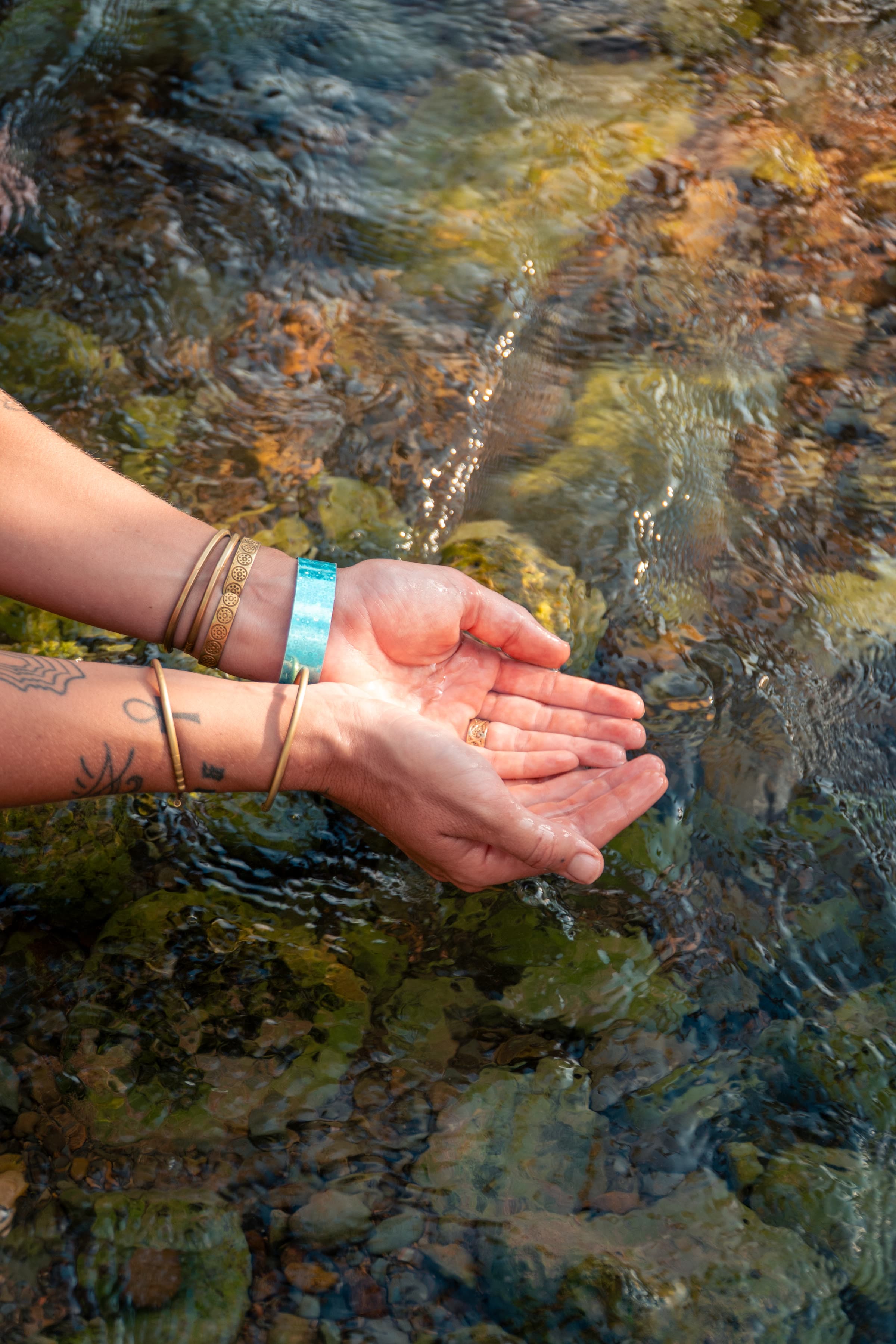 Cupped tattooed hands reach into a clear rocky stream, gold bangles and a wide turquoise cuff catching the light.